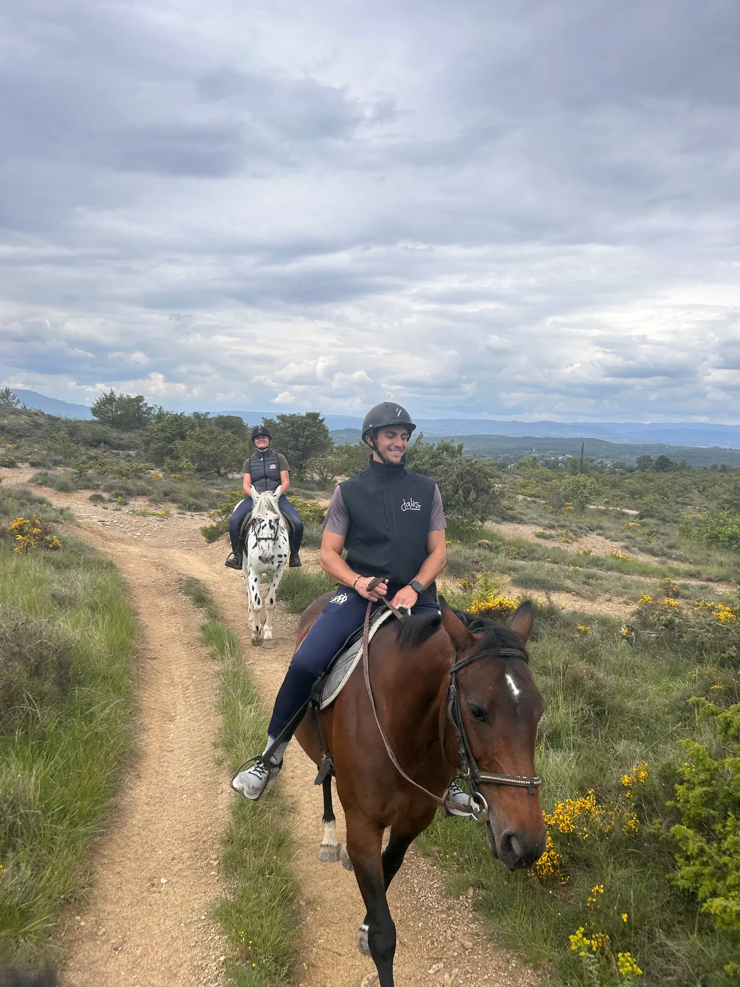 Deux cavaliers avançant sur un chemin de garrigue avec les collines en arrière-plan